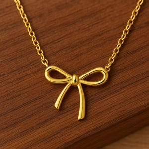 May include: A gold-colored necklace featuring a bow pendant. The bow is a delicate, three-dimensional design with curved lines and a small knot in the center. The chain is a fine link style, and the necklace is displayed on a wooden surface.