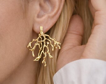 May include: Gold-colored coral-shaped earrings. The earrings are worn on a person's ear. The earrings have a textured surface and a branch-like design. The earrings are a bright gold color.