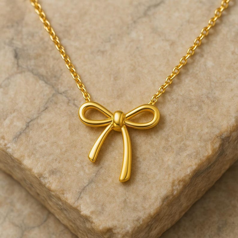 May include: A gold-colored necklace featuring a bow pendant. The bow is centered on a delicate chain, with the bow's loops and tails clearly defined. The necklace is displayed on a textured, light brown surface.