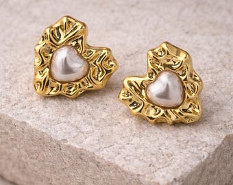 May include: A pair of gold-colored heart-shaped stud earrings. Each earring features a pearl-like center and a textured, ornate gold border. The earrings are displayed on a light-colored, textured surface.