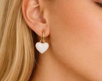 May include: Gold hoop earrings with a white heart pendant. The earrings are a simple, elegant design, featuring a polished gold finish and a smooth, heart-shaped charm. The heart pendant is a solid white color.