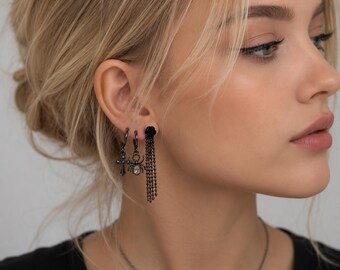 May include: A set of three black earrings featuring a cross charm, a spider charm with a clear crystal center, and a black rose stud with a long, dangling multi-strand chain. The earrings are made of dark metal and have a polished finish.