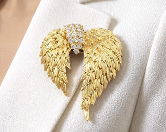 May include: A gold-colored angel wing brooch with a central cluster of clear crystals. The wings have a textured, feather-like design. The brooch is pinned to a white blazer.