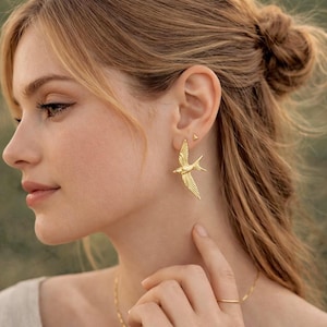 May include: Gold-colored bird earrings. The earrings are in the shape of a bird in flight, with detailed wing and tail feathers. A small gold stud earring is also visible. The jewelry is worn on a person.