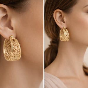 May include: Gold-colored filigree hoop earrings. The earrings have an oval shape with intricate openwork designs. The earrings are worn by a person with light skin and brown hair. The earrings are a classic jewelry piece.