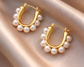 May include: A pair of gold-colored hoop earrings, each adorned with a semi-circle of small, white pearls. The earrings are set against a soft, satin-like background, creating a luxurious aesthetic.