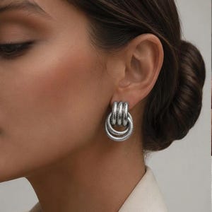 May include: A silver-toned earring featuring a triple-loop design with a textured, cable-like finish. The earring consists of three interconnected circular segments that hang vertically from the earlobe, creating a layered, metallic appearance.