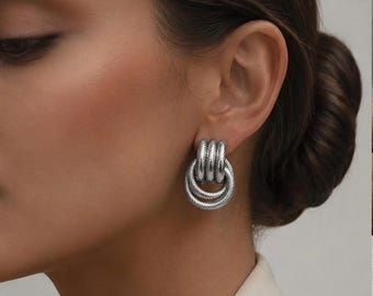 May include: A silver-toned earring featuring a triple-loop design with a textured, cable-like finish. The earring consists of three interconnected circular segments that hang vertically from the earlobe, creating a layered, metallic appearance.