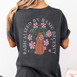 May include: A dark gray t-shirt with a floral design and the text "RADICALIZED BY BASIC DECENCY" in a semi-circular arch. The design features a brown vase with pink flowers. The word "Pepper" is printed at the bottom.