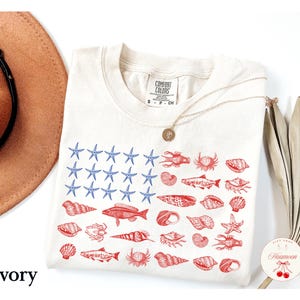 May include: An ivory short-sleeve t-shirt featuring a graphic print arranged like a flag. The left section displays blue starfish, while the right section features red illustrations of various fish, crabs, and seashells. The shirt has a crew neckline.