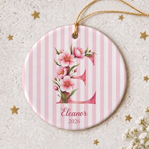 May include: A round ceramic ornament featuring a pink and white vertical striped background. The letter E is printed in a large pink font, decorated with pink floral illustrations. The name Eleanor and the year 2026 are printed below.