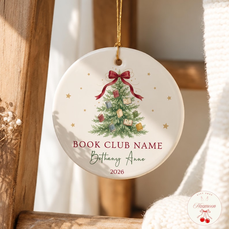 May include: A round white ceramic ornament hanging by a gold string. The center features a green Christmas tree decorated with small books and a red bow at the top. Text reads BOOK CLUB NAME, Bethany Anne, and 2026, surrounded by gold stars.