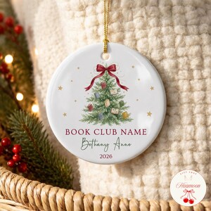 May include: A round white ceramic ornament featuring a green Christmas tree decorated with small books, topped with a red ribbon bow. The text reads BOOK CLUB NAME, Bethany Anne, and 2026. A gold string is attached to the top for hanging.