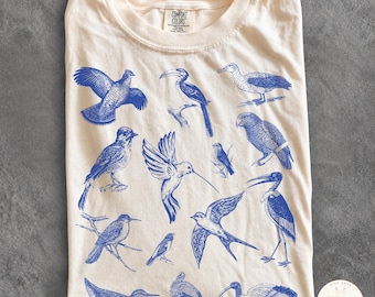 Puede incluir: Una camiseta de color marfil con una colección de ilustraciones en tinta azul de varios pájaros, incluyendo un colibrí, un tucán y una cigüeña. La camiseta tiene un cuello redondo y una etiqueta de la marca Comfort Colors en el interior.