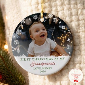 May include: A round white ceramic ornament featuring a photo of a baby in a white shirt. The design includes white snowflakes and gift icons, with the text: FIRST CHRISTMAS AS MY Grandparents LOVE, HENRY 2026. It hangs from a gold cord.