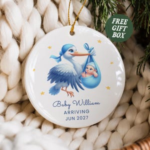 May include: A round white ceramic ornament featuring a blue stork carrying a baby in a blue bundle. The text reads Baby William ARRIVING JUN 2027. The ornament has a gold hanging string and is decorated with small yellow stars.