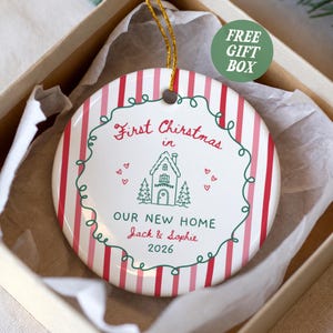 May include: A round ceramic ornament with red and pink vertical stripes, featuring a green line-art wreath, a house illustration, and the text First Christmas in OUR NEW HOME Jack & Sophie 2026, suspended by a gold string inside a gift box.