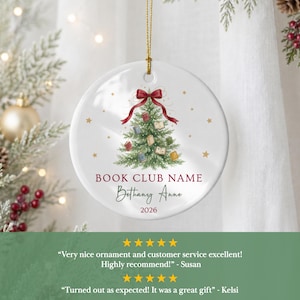 May include: A round white ceramic ornament featuring a green Christmas tree decorated with small books and a red ribbon bow at the top. The text reads BOOK CLUB NAME, Bethany Anne, and 2026 in red and black script, suspended by a gold string.