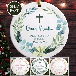 May include: A white ceramic Christmas ornament with a cross and floral wreath design. The ornament features the name "Owen Brooks", the word "DEDICATED", a date, and a Bible verse. The text "SHIP FAST" is in a circle.