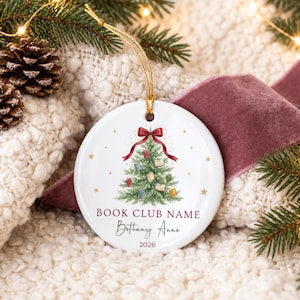 May include: A round white ceramic ornament featuring a green Christmas tree decorated with small books and a red bow at the top. The text reads BOOK CLUB NAME, Bethany Anne, and 2026. A gold string is attached to the top for hanging purposes.