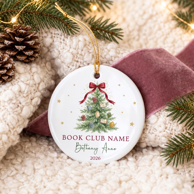 May include: A round white ceramic ornament featuring a green Christmas tree decorated with small books and a red bow at the top. The text reads BOOK CLUB NAME, Bethany Anne, and 2026. A gold string is attached to the top for hanging purposes.