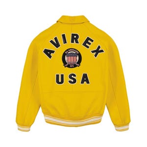 May include: A bright yellow varsity jacket featuring the text AVIREX in black arched lettering across the upper back. Below the text is a circular patch with a flag design and the words ALL AMERICANS ESTABLISHED 1975. The word USA is printed in large black letters below the patch, with white and yellow striped ribbed cuffs and hem.