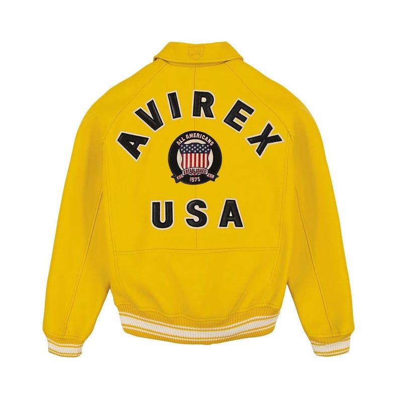 May include: A bright yellow varsity jacket featuring the text AVIREX in black arched lettering across the upper back. Below the text is a circular patch with a flag design and the words ALL AMERICANS ESTABLISHED 1975. The word USA is printed in large black letters below the patch, with white and yellow striped ribbed cuffs and hem.