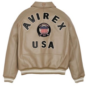 May include: A tan leather bomber jacket featuring a ribbed collar, cuffs, and hem with white stripes. The back displays the black embroidered text AVIREX and USA, along with a circular patch featuring a flag design and the text ALL AMERICANS.