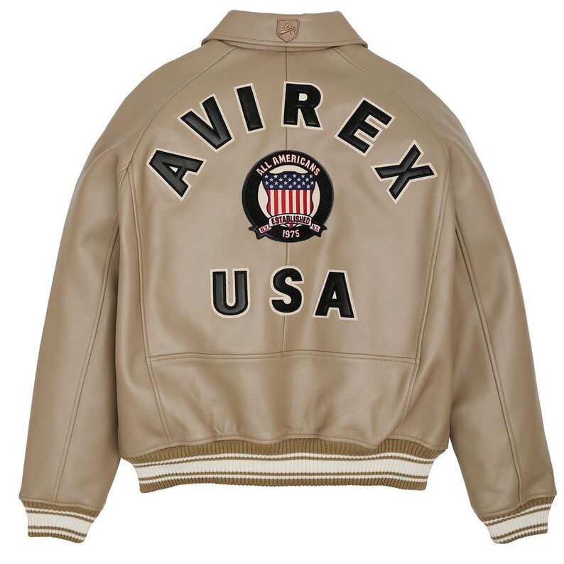 May include: A tan leather bomber jacket featuring a ribbed collar, cuffs, and hem with white stripes. The back displays the black embroidered text AVIREX and USA, along with a circular patch featuring a flag design and the text ALL AMERICANS.
