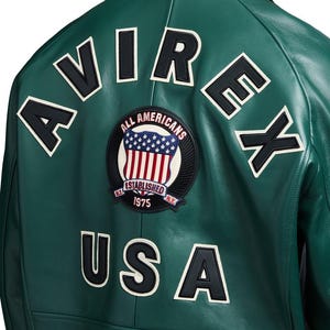 May include: A dark green leather jacket featuring the word AVIREX arched across the upper back in black and white lettering. Below this, a circular patch displays a shield with stars and stripes, the text ALL AMERICANS, ESTABLISHED 1975, and USA.