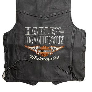 Könnte beinhalten: Schwarze Motorradweste aus Leder mit dem Harley-Davidson-Logo in Weiß und Orange. Die Weste hat ein Schnürdesign an den Seiten und das Wort "Motorcycles" steht unter dem Logo.