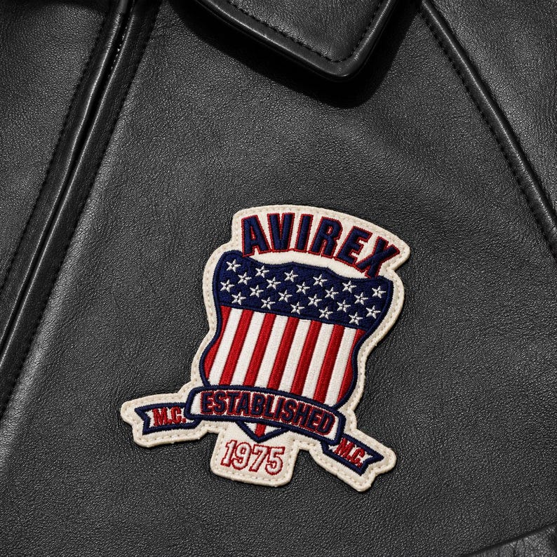 May include: A black leather jacket featuring an embroidered patch. The patch displays a shield with red and white stripes and white stars on a blue field, with the text AVIREX, ESTABLISHED 1975, and M.C. in red, white, and blue thread.