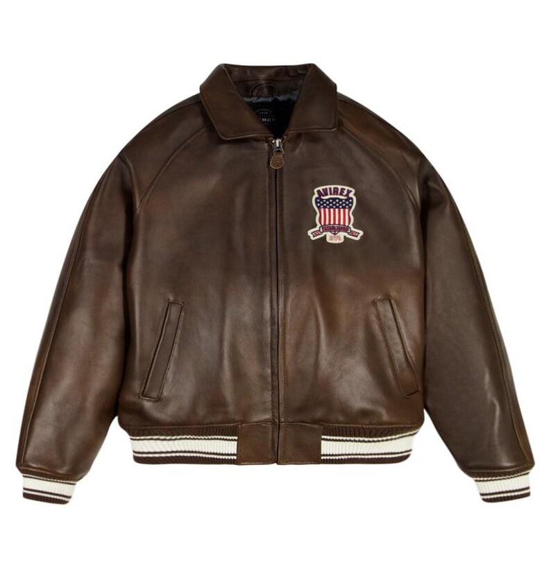 May include: A dark brown leather jacket with a front zipper closure, a pointed collar, and two side pockets. The chest features an embroidered patch with the text AVIREX ESTABLISHED 1975 and a red, white, and blue flag design. The cuffs and hem are ribbed.