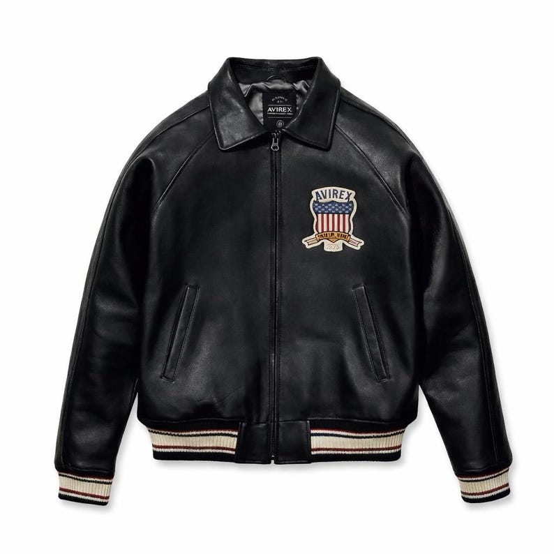 May include: A black leather jacket featuring a front zipper closure, a classic collar, and two side pockets. On the left chest, there is a patch displaying the text AVIREX above a shield with a red, white, and blue flag design. The cuffs and hem are ribbed with black, white, and red stripes.
