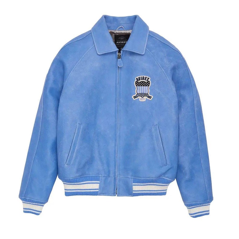 May include: A light blue jacket with a full-length front zipper, a pointed collar, and long sleeves. The garment features a white and blue striped ribbed hem and cuffs, two side pockets, and an embroidered patch on the chest reading AVIREX.