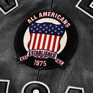 May include: A black leather jacket featuring a circular embroidered patch. The patch displays the text ALL AMERICANS, ESTABLISHED 1975, and M.C. surrounding a shield with a blue field of white stars above red and white vertical stripes.