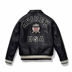 May include: A black leather jacket featuring the word AVIREX in white lettering across the upper back. Below the text is a circular patch with an American flag design and the text BALL AMERICAN EST 1975. The word USA is printed in white below.