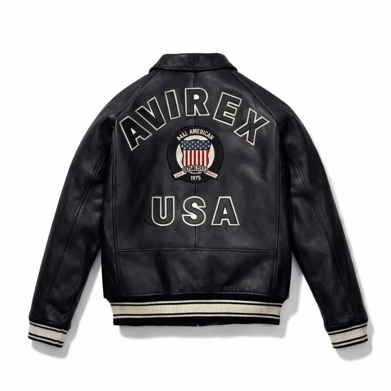 May include: A black leather jacket featuring the word AVIREX in white lettering across the upper back. Below the text is a circular patch with an American flag design and the text BALL AMERICAN EST 1975. The word USA is printed in white below.