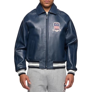 May include: A navy blue leather bomber jacket featuring a full-length front zipper, a classic collar, and ribbed cuffs and hem with white stripes. An embroidered patch on the chest displays the text AVIREX with a red, white, and blue flag design.