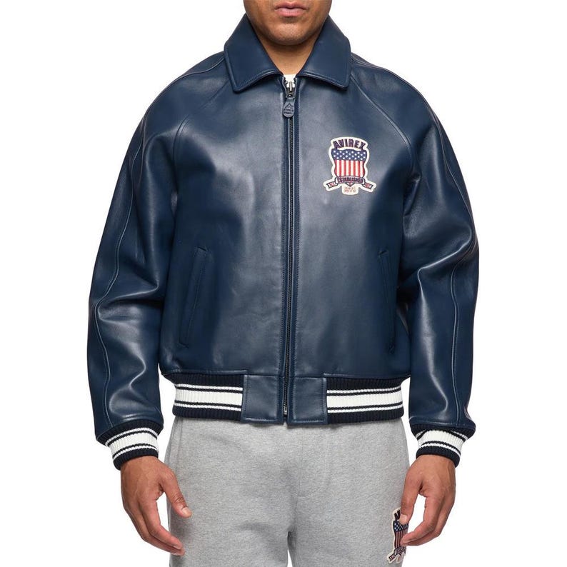 May include: A navy blue leather bomber jacket featuring a full-length front zipper, a classic collar, and ribbed cuffs and hem with white stripes. An embroidered patch on the chest displays the text AVIREX with a red, white, and blue flag design.