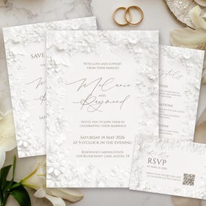 May include: Elegant white wedding invitation suite with floral accents. Includes invitation, RSVP, and details cards. Features the names "Melanie and Raymond" and wedding date, May 14, 2026. Gold wedding rings and white flowers complete the arrangement.
