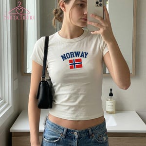 Può includere: Una t-shirt a maniche corte e girocollo color bianco sporco con la scritta NORWAY in grassetto blu sopra una bandiera norvegese rossa, bianca e blu centrata sul petto. Il capo è abbinato a una borsa a tracolla nera e a un paio di jeans blu.