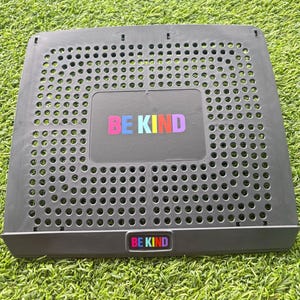 Könnte beinhalten: Ein dunkelgrauer Laptop-Schreibtisch aus Kunststoff mit perforierter Oberfläche. Der Schreibtisch trägt die Worte "BE KIND" in bunten Buchstaben oben und unten. Der Schreibtisch steht auf einer grünen, grasbewachsenen Oberfläche.
