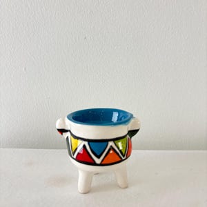 Peut inclure: Petit pot en céramique blanche avec intérieur bleu et trois petits pieds. Le pot présente un motif géométrique coloré avec des triangles rouges, jaunes, bleus et oranges. Il possède deux petites poignées de chaque côté.