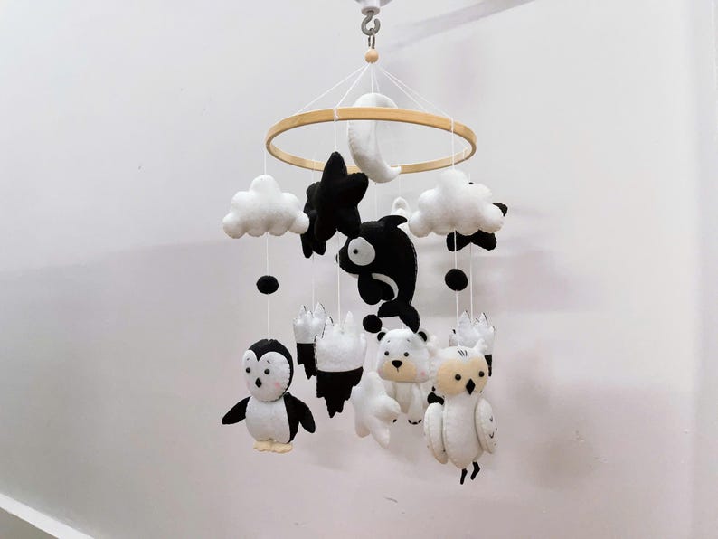 K&ouml;nnte beinhalten: Ein Baby-Mobile mit Filz-Tier- und Himmelsk&ouml;rperformen in Schwarz, Wei&szlig; und Creme. Das Mobile enth&auml;lt einen Pinguin, Eisb&auml;ren, eine Eule, Wolken, Sterne und einen Mond, die an einem Holzreifen aufgeh&auml;ngt sind.