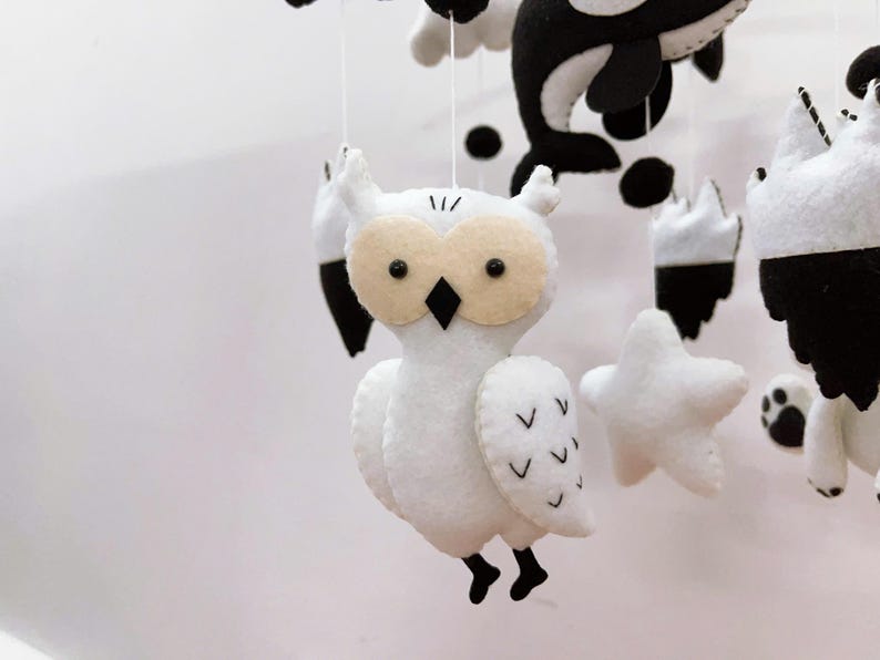 K&ouml;nnte beinhalten: Handgefertigtes Babymobile mit Filztierformen in Wei&szlig;, Schwarz und Beige. Das Mobile enth&auml;lt eine Eule, einen Orca und Sterne. Die Eule hat beige Augen und einen schwarzen Schnabel. Das Mobile ist an wei&szlig;en Schn&uuml;ren aufgeh&auml;ngt.