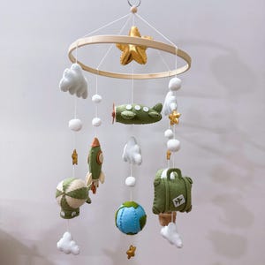 Puede incluir: Un m&oacute;vil colgante para habitaci&oacute;n infantil con marco circular de madera y adornos de fieltro que incluyen una estrella dorada, un avi&oacute;n verde, un cohete, un globo aerost&aacute;tico, un globo terr&aacute;queo, una maleta con icono de avi&oacute;n y nubes blancas.