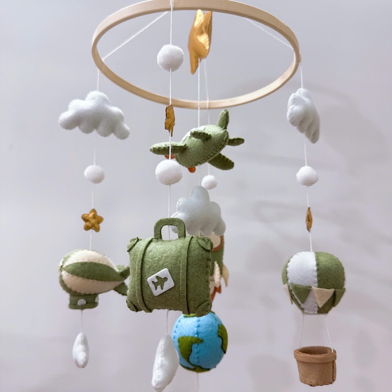 Puede incluir: Un m&oacute;vil colgante para guarder&iacute;a con marco circular de madera y adornos de fieltro que incluyen un avi&oacute;n verde, una maleta verde con icono de avi&oacute;n, un globo terr&aacute;queo azul y verde, un globo aerost&aacute;tico, un dirigible, nubes blancas y estrellas doradas.