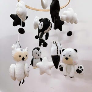 K&ouml;nnte beinhalten: Ein Baby-Mobile mit Tierfiguren aus Filz in Schwarz, Wei&szlig; und Beige. Das Mobile umfasst eine Eule, einen Pinguin, einen B&auml;ren, einen Orca, Sterne, Wolken und abstrakte Formen, die an einem Holzreifen aufgeh&auml;ngt sind.