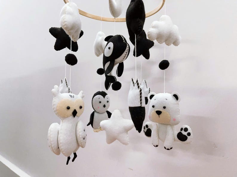 K&ouml;nnte beinhalten: Ein Baby-Mobile mit Tierfiguren aus Filz in Schwarz, Wei&szlig; und Beige. Das Mobile umfasst eine Eule, einen Pinguin, einen B&auml;ren, einen Orca, Sterne, Wolken und abstrakte Formen, die an einem Holzreifen aufgeh&auml;ngt sind.